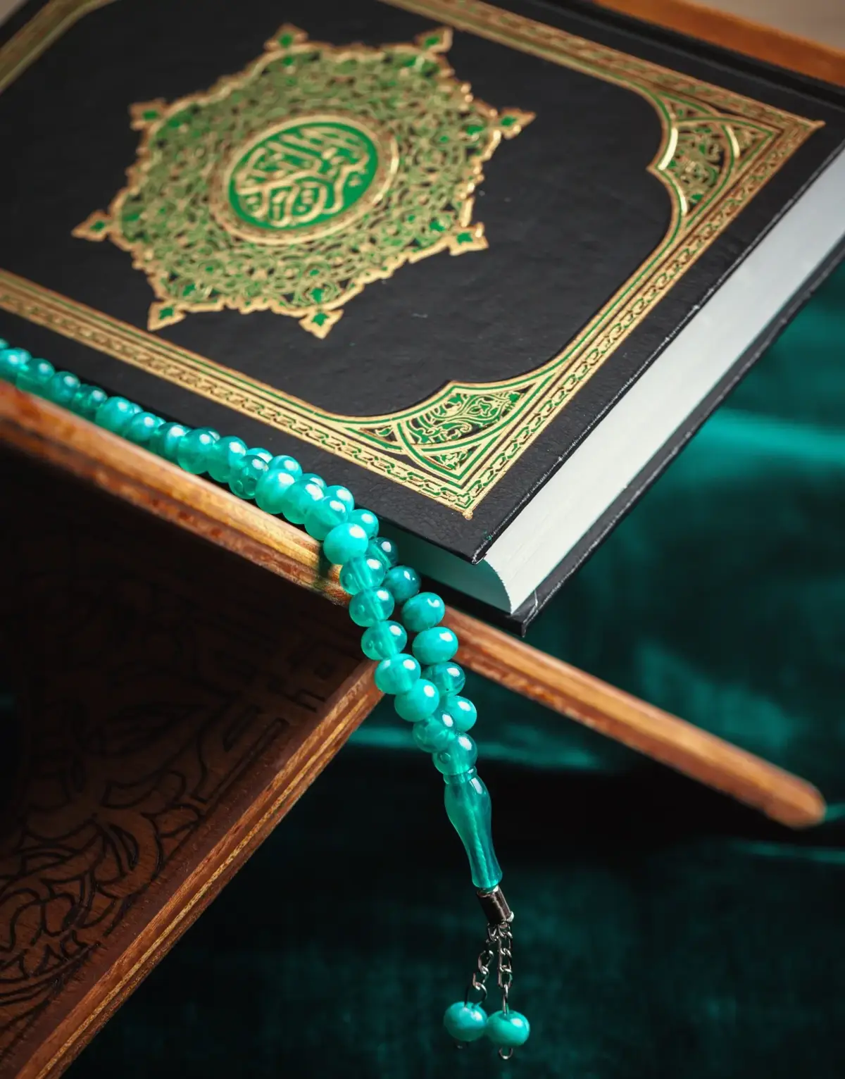 A close-up shot of the Holy Quran featuring a black cover with intricate gold and green embossing, resting on a carved wooden book stand with a vibrant string of turquoise prayer beads draped over it against a dark green velvet backdrop.