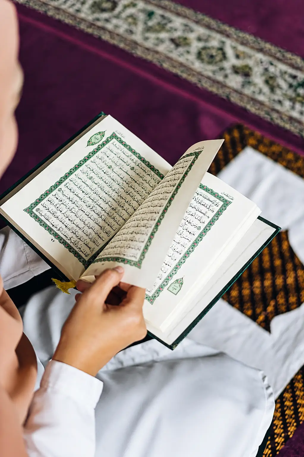 An over-the-shoulder close-up view of an individual wearing a white shirt, sitting on a purple prayer rug, and turning the pages of an open Holy Quran, chosen for the website's About Us section.